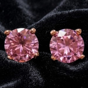 May include: A pair of round-cut pink stud earrings featuring faceted stones secured by four metal prongs. The earrings display a vibrant pink hue with geometric light reflections across the surface of each circular gemstone setting.