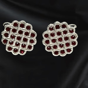 May include: A pair of silver-toned cufflinks with a woven design. Each cufflink features a floral pattern with small, round, deep red gemstones set within the woven silver. The cufflinks are displayed against a dark background.