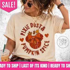 May include: A cream-colored short-sleeve t-shirt featuring a graphic of a pumpkin with a bow and autumn leaves. The text reads Pixie Dust & Pumpkin Spice. The design includes two small hearts and has a distressed, textured appearance.