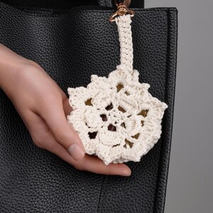 May include: A cream-colored, crocheted flower-shaped bag charm with a woven strap and a gold-tone clasp. The charm is held in front of a black leather tote bag.