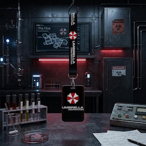Può includere: Laccetto nero con il logo e il testo Umbrella Corporation. È appeso in un laboratorio con provette, uno schermo di computer con un diagramma e segnali di pericolo biologico. Il porta badge mostra anche il logo Umbrella Corporation.