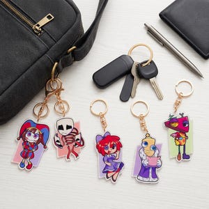 Puede incluir: Una colección de llaveros acrílicos con personajes de dibujos animados coloridos. Cada llavero tiene un anillo dorado y un diseño único, que incluye un bufón, un esqueleto y un robot. Los llaveros se muestran con llaves, un bolígrafo y una bolsa negra.