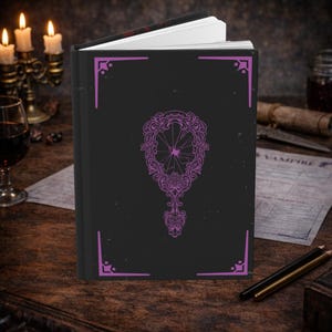 Puede incluir: Un diario negro con un diseño de espejo morado adornado en la portada. El diario tiene un borde decorativo morado y está abierto para revelar páginas blancas. La imagen está sobre una mesa de madera con velas y una copa de vino.