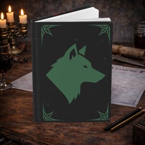 Puede incluir: Un diario de tapa dura con una cubierta negra que presenta una silueta de cabeza de lobo verde. La cubierta tiene diseños decorativos en las esquinas y una textura desgastada. El diario está abierto, revelando páginas blancas, sobre una superficie de madera.