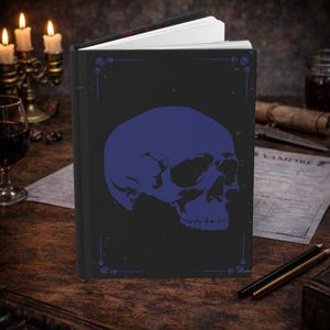 Puede incluir: Un diario negro con un diseño de calavera azul en la portada. La calavera está centrada y detallada, con elementos decorativos en las esquinas. El diario está abierto, revelando páginas blancas, sobre una superficie de madera.