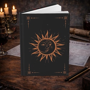 Puede incluir: Diario negro con un diseño de sol y luna naranja en la portada. El sol y la luna se besan. El diario está abierto, revelando páginas blancas. La portada tiene detalles decorativos de estrellas y líneas. El diario está sobre una superficie de madera.