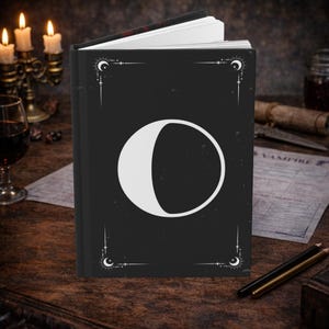 Puede incluir: Un diario negro con un diseño de media luna blanca en la portada. El diario tiene detalles decorativos en las esquinas y un lomo a juego. La palabra "VAMPIRE" es visible en un papel en el fondo.
