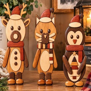 Könnte beinhalten: Drei Holzfiguren in Form eines Rentiers, einer Katze und eines Pinguins, die jeweils eine rote Weihnachtsmütze und einen Schal tragen. Das Rentier hat ein Geweih, die Katze Schnurrhaare und der Pinguin hält einen kleinen Baum. Gefertigt aus Holz.