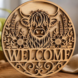 May include: A circular wooden wall sign featuring a laser-cut design of a Highland cow head centered above the word WELCOME. The artwork includes wheat stalks and sunflower motifs on both sides, with decorative scrollwork at the bottom edge.