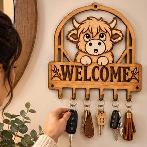以下が含まれることがあります： アーチ型のトップ、ハイランド牛の漫画イラスト、中央にWELCOMEの文字が刻印された、ライトブラウンの木製壁掛けキーホルダー。5つの金属製フックがあり、それぞれに様々なキーホルダーが付いた鍵のセットが掛けられています。
