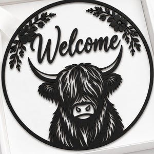 Könnte beinhalten: Ein rundes, schwarzes Wandschild mit einer Highland-Rind-Illustration und dem Wort Welcome in Schreibschrift, umrahmt von floralen Akzenten. Das Schild liegt in einer weißen Geschenkbox mit weißem Band, einem Anhänger und einer Karte.