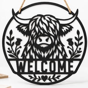 Könnte beinhalten: Ein kreisförmiger schwarzer Wandbehang mit einem lasergeschnittenen Design eines Hochlandrindes mit langen Hörnern und zotteligem Fell. Das Wort WELCOME ist unten in fetten Großbuchstaben ausgeschnitten, flankiert von Blumenzweigen und einem kleinen Herz.