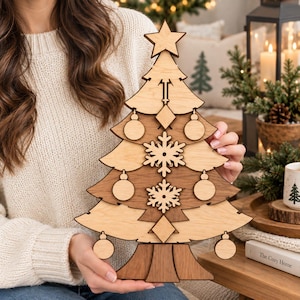 May include: A layered wooden Christmas tree decoration featuring alternating light and dark wood tones. The tree is adorned with cut-out ornaments including circular baubles, two large snowflakes, diamond shapes, and a star topper at the peak.