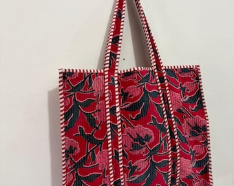 May include: A quilted tote bag featuring a vibrant red background with a dark green and pink floral pattern. The bag includes two long shoulder straps and edges finished with a white and red striped fabric trim, creating a structured look.