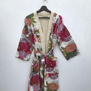 Pode incluir: Um roupão longo acolchoado com um padrão floral em tons de rosa, laranja, verde e branco. O roupão tem um interior creme e um cinto a condizer. O roupão está pendurado num cabide de madeira.