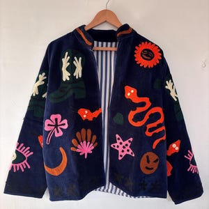 May include: A navy blue jacket adorned with vibrant embroidered motifs. The jacket showcases embroidered designs such as a sun, snake, stars, and flowers. It has a striped lining and is suspended from a wooden hanger.