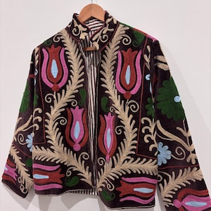 May include: A brown velvet jacket with vibrant floral embroidery. The jacket has a high collar and a striped lining. The embroidery features red, pink, blue, and green flowers with gold accents. The jacket is displayed on a wooden hanger.