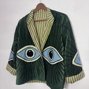May include: A green velvet jacket with embroidered eye designs. The jacket has a striped lining and cuffs with a green, white, and pink pattern. The eye designs are blue, black, and cream coloured.