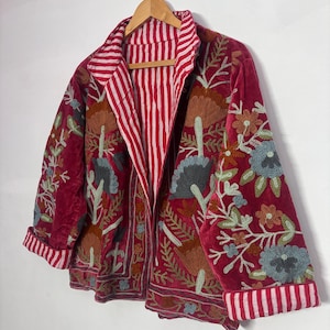 May include: A burgundy velvet jacket with floral embroidery and a striped red and white lining. The jacket is hanging on a wooden hanger. The cuffs and collar are also striped.