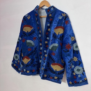 Pode incluir: Casaco de veludo azul com bordados florais em vermelho, amarelo e azul. O casaco tem gola mandarim e mangas três quartos. O forro é listrado. O casaco está pendurado em um cabide de madeira.