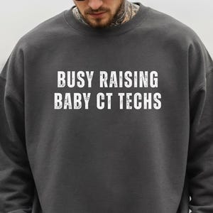 May include: A dark gray sweatshirt with the text "BUSY RAISING BABY CT TECHS" in white. The sweatshirt is a crew neck style and is being worn by a person wearing sunglasses.