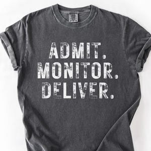 May include: A charcoal gray short-sleeve t-shirt with a distressed white graphic print on the chest. The text reads ADMIT. MONITOR. DELIVER. in a bold, sans-serif font. The garment features a crew neckline and a vintage-style washed finish.
