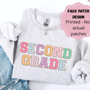 May include: A light gray crewneck sweatshirt featuring the text SECOND GRADE in multicolored varsity-style letters with a faux patch design. The text is printed in pink, blue, purple, and orange with a gold outline. A pink label states FAUX PATCH DESIGN Printed - Not actual patches.
