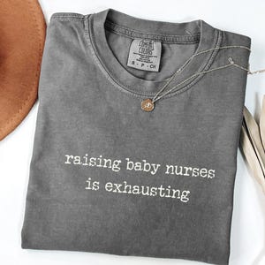 May include: A grey Comfort Colors t-shirt with the text "raising baby nurses is exhausting" in white. A silver necklace with a small pendant is draped across the shirt. A brown hat is visible in the upper left corner.