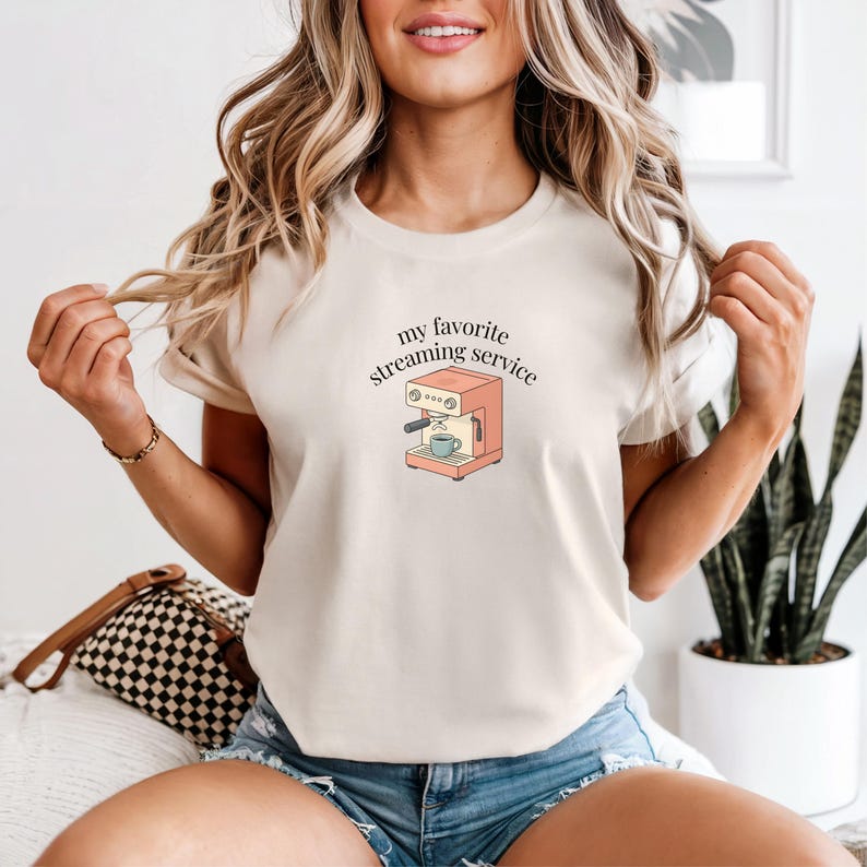 Close-up of a woman wearing a light ivory t-shirt featuring the phrase “my favorite streaming service” arched above a coral espresso machine graphic, styled with denim shorts in a bright, cozy indoor setting.