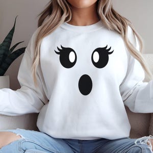 May include: White sweatshirt featuring a surprised ghost face design. The ghost face has large black eyes with white pupils and black eyelashes, and a black oval mouth. The sweatshirt is a casual, comfortable style.