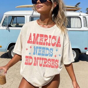 May include: Cream-colored t-shirt with the text "AMERICA NEEDS ED NURSES" in red and blue. The shirt has a relaxed fit and short sleeves. A light blue van with a surfboard on top is in the background.