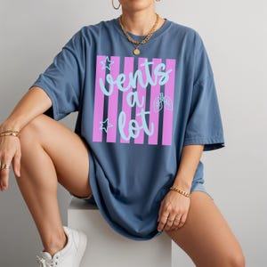 Puede incluir: Camiseta azul con la frase "Vents a lot" en escritura azul claro, sobre un fondo de rayas rosas. El dise&ntilde;o incluye un gr&aacute;fico de pulmones y estrellas. La camiseta es de talla grande.