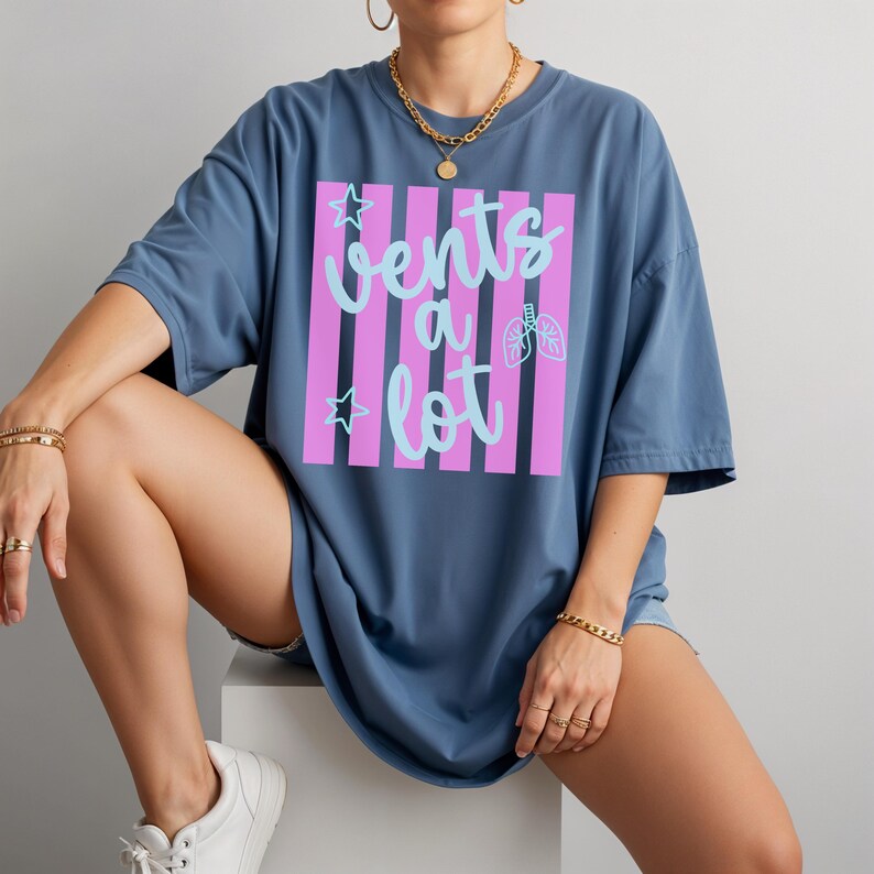 Puede incluir: Camiseta azul con la frase "Vents a lot" en escritura azul claro, sobre un fondo de rayas rosas. El dise&ntilde;o incluye un gr&aacute;fico de pulmones y estrellas. La camiseta es de talla grande.