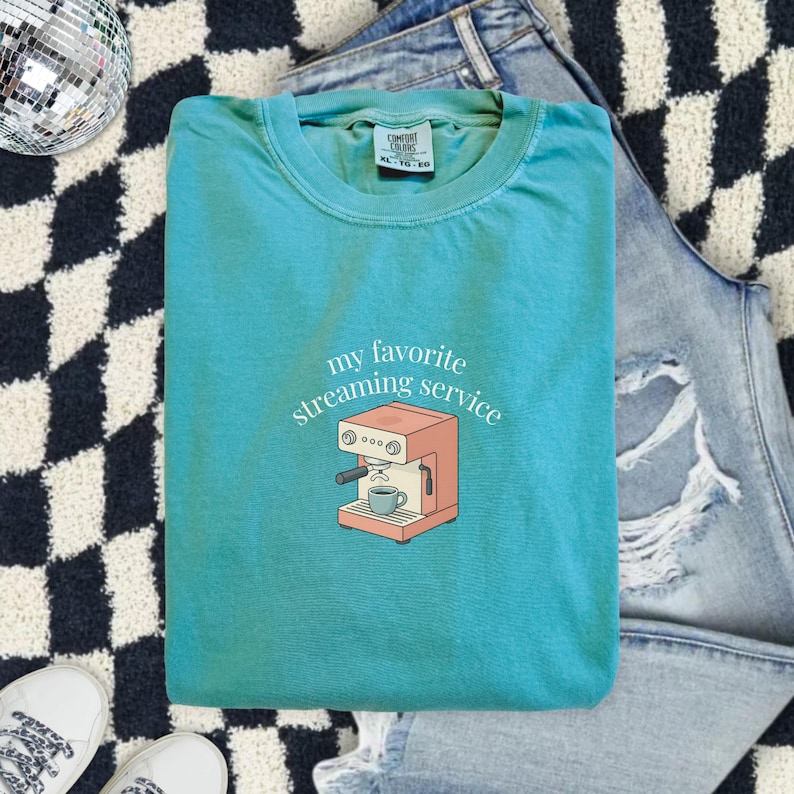 Folded teal Comfort Colors t-shirt with minimalist espresso machine graphic and “my favorite streaming service” text, styled flat lay with denim jeans and sneakers for an aesthetic coffee lover outfit.