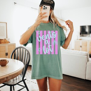 Puede incluir: Camiseta verde salvia con el texto "Vents a lot" en escritura azul claro, sobre un fondo de rayas rosas. El dise&ntilde;o incluye estrellas y un gr&aacute;fico de pulmones. La camiseta es de estilo casual y unisex.