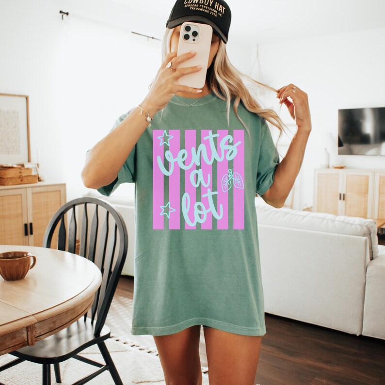 Puede incluir: Camiseta verde salvia con el texto "Vents a lot" en escritura azul claro, sobre un fondo de rayas rosas. El dise&ntilde;o incluye estrellas y un gr&aacute;fico de pulmones. La camiseta es de estilo casual y unisex.