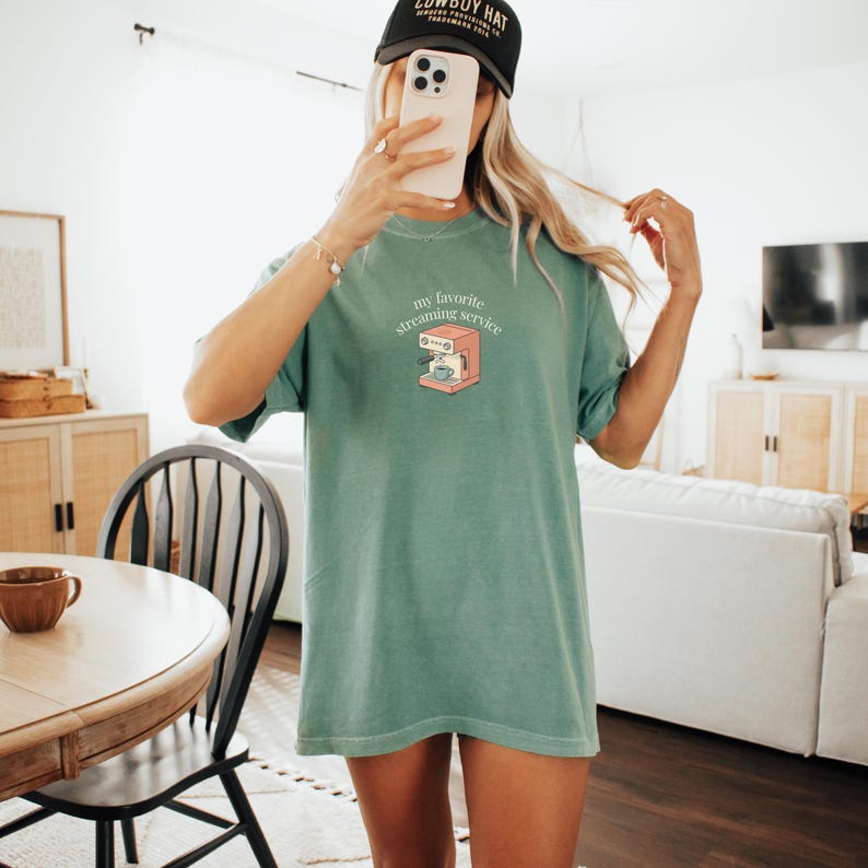 May include: A seafoam green t-shirt with the phrase "my favorite streaming service" and a coffee machine graphic. The shirt is a relaxed fit and has short sleeves. A black baseball cap is worn.