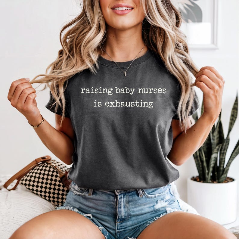 May include: Dark grey t-shirt with the white text "raising baby nurses is exhausting". The t-shirt is a crew neck style and is worn with denim shorts. The image is taken in natural light.