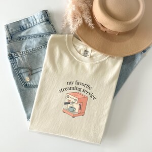 May include: Cream-colored t-shirt with the text "my favorite streaming service" and a graphic of a coffee machine. The shirt is paired with light blue jeans, a gold necklace, and a tan hat.