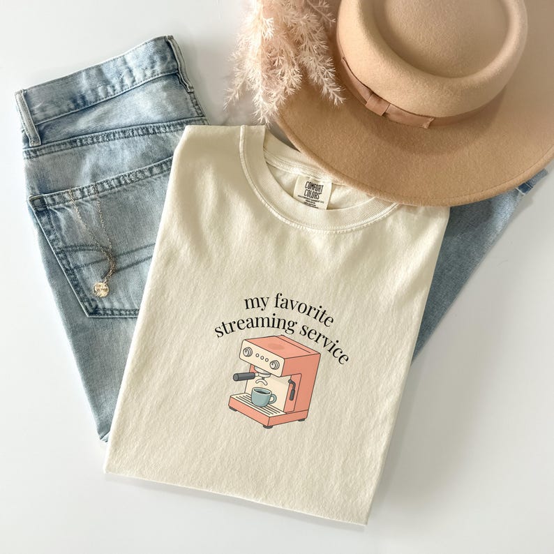 May include: Cream-colored t-shirt with the text "my favorite streaming service" and a graphic of a coffee machine. The shirt is paired with light blue jeans, a gold necklace, and a tan hat.