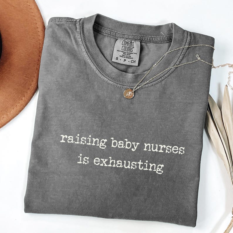 May include: A grey Comfort Colors t-shirt with the text "raising baby nurses is exhausting" in white. A silver necklace with a small pendant is draped across the shirt. A brown hat is visible in the upper left corner.