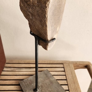 Pode incluir: Um fragmento fossilizado de osso de saurópode, com uma superfície rugosa, texturizada e de cor bege. O espécime está montado num suporte de metal preto com uma base quadrada, que inclui uma pequena etiqueta com texto identificativo.