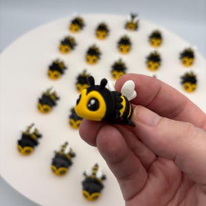 May include: A collection of small, cartoon-style bee figurines featuring black and yellow striped bodies, white wings, and large eyes. One bee is held in the foreground, while numerous identical bee figures are arranged on a white surface.