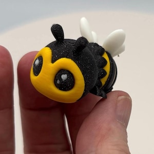 May include: A 3D-printed bumblebee figurine with a black and yellow body, large eyes, and white wings. The bee has a cute, cartoonish design. The figurine is held between two fingers.