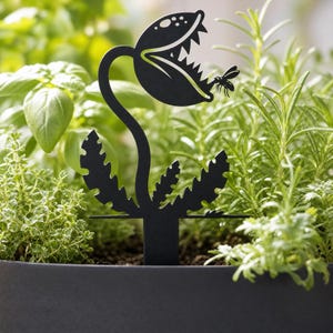 May include: A black metal plant stake featuring a silhouette of a carnivorous plant with an open mouth and sharp teeth, capturing a small insect. The design includes a curved stem and jagged leaves, with a flat base for insertion into soil.