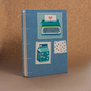 May include: A blue fabric-covered journal with exposed white stitching along the spine. The cover features three stitched patches: a teal typewriter with a red heart, a blue milk carton with white flowers, and a white square with black stars.