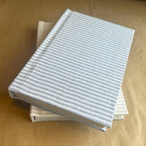 May include: Two hardcover journals with striped covers. The top journal has a blue and white striped pattern, while the bottom journal has a beige and white striped pattern. The journals are stacked on top of each other.