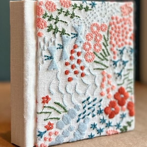 Könnte beinhalten: Quadratische, cremefarbene Leinwand mit aufwendiger Blumenstickerei. Die Stickerei zeigt verschiedene Stiche in Korallen-, Hellblau- und Grüntönen, wodurch ein strukturiertes, künstlerisches Design entsteht. Die Leinwand ist auf einem dicken, weißen Rahmen montiert.