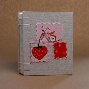 May include: A light grey linen-covered journal with exposed white thread binding. The front cover features three stitched fabric patches: a pink square with a bicycle illustration, a cream square with a red strawberry, and a red square with gold stars.