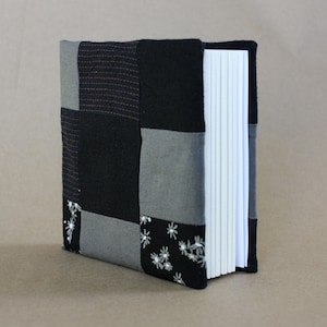 May include: A handcrafted journal with a patchwork cover in black and grey tones. The cover showcases diverse fabric textures and patterns, including floral embroidery. The journal has white pages and a black fabric spine.